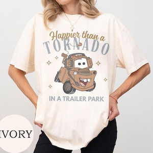 May include: Ivory-colored t-shirt featuring a cartoon image of Mater from the movie Cars. The shirt has the text "Happier than a Tornado in a Trailer Park" in brown and gray. The shirt is a short-sleeved, casual style.
