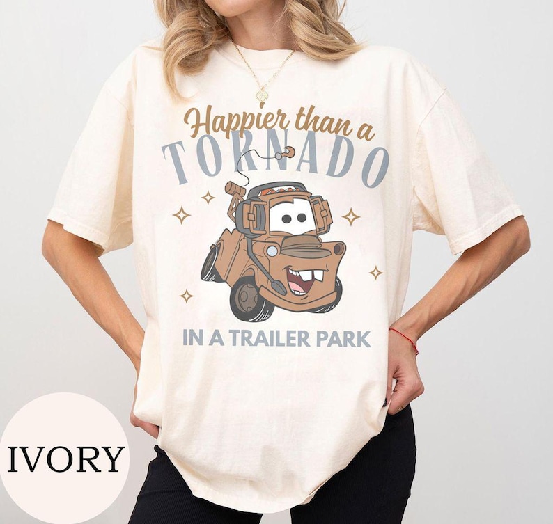 May include: Ivory-colored t-shirt featuring a cartoon image of Mater from the movie Cars. The shirt has the text "Happier than a Tornado in a Trailer Park" in brown and gray. The shirt is a short-sleeved, casual style.