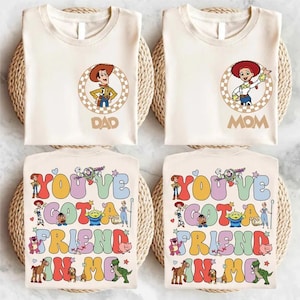 May include: Four cream-coloured t-shirts. Two shirts feature a circular graphic with a cartoon cowboy and the word "DAD". The other two shirts have a circular graphic with a cartoon cowgirl and the word "MOM". The bottom shirts have the text "YOU'VE GOT A FRIEND IN ME".