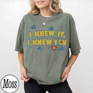 May include: An olive green short-sleeve t-shirt featuring the yellow text I KNEW IT, I KNEW YOU with a red heart containing the letters TS and a blue ball graphic. The design includes small blue stars and cloud shapes printed on the front.