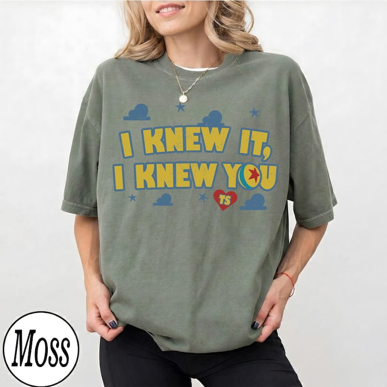 May include: An olive green short-sleeve t-shirt featuring the yellow text I KNEW IT, I KNEW YOU with a red heart containing the letters TS and a blue ball graphic. The design includes small blue stars and cloud shapes printed on the front.