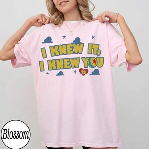 May include: A light pink short-sleeved t-shirt featuring the yellow text I KNEW IT, I KNEW YOU with a red heart containing the letters TS, surrounded by blue cloud and star graphics. The design has a distressed, vintage-inspired texture.