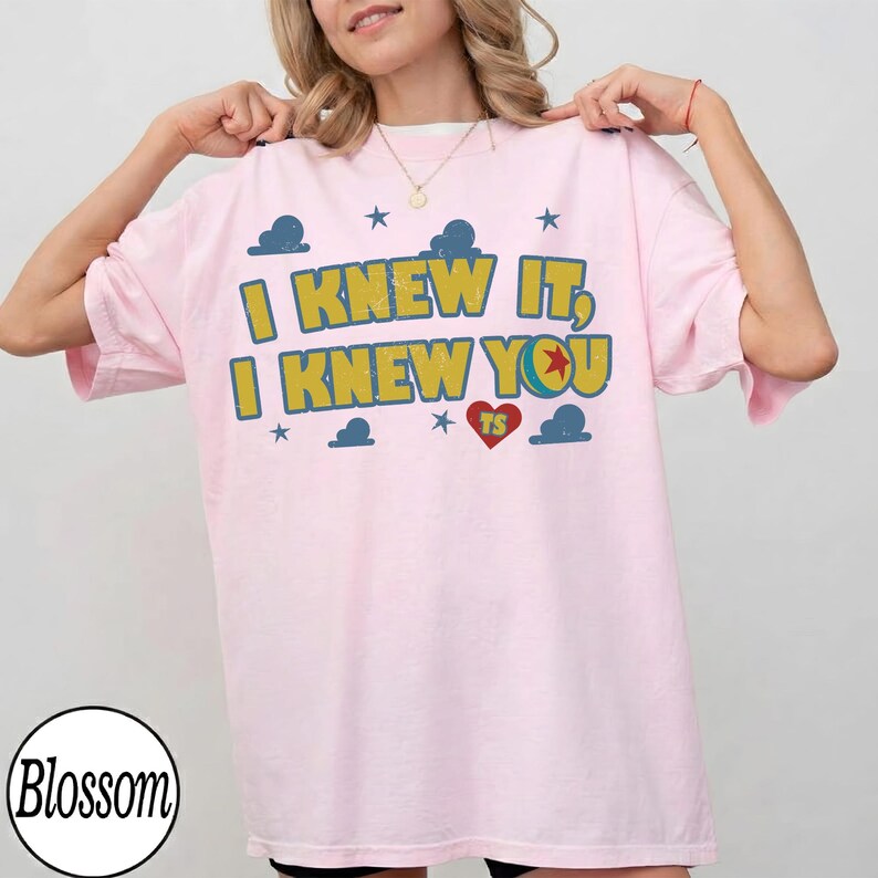 May include: A light pink short-sleeved t-shirt featuring the yellow text I KNEW IT, I KNEW YOU with a red heart containing the letters TS, surrounded by blue cloud and star graphics. The design has a distressed, vintage-inspired texture.