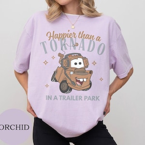 May include: Orchid-colored t-shirt featuring a cartoon image of Tow Mater from the movie Cars. The shirt has the text "Happier than a Tornado in a Trailer Park" in brown and gray. The design includes star accents.