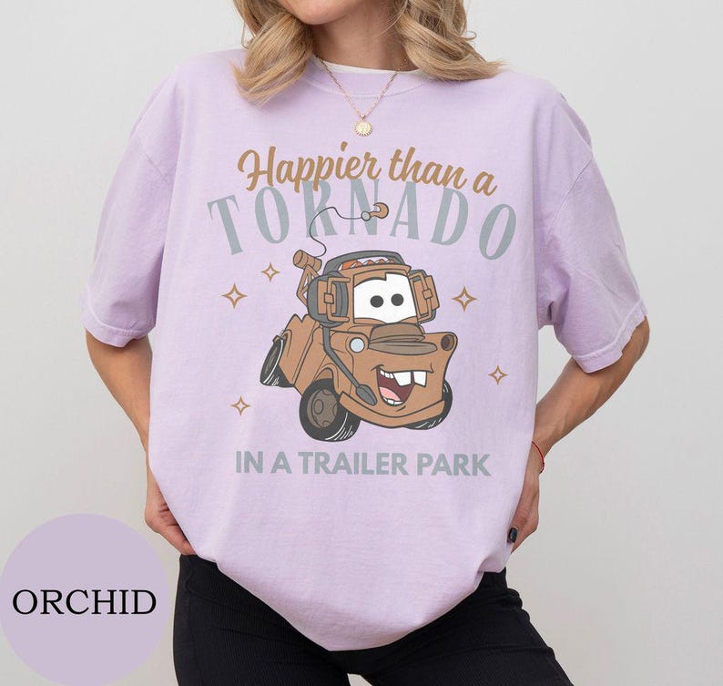 May include: Orchid-colored t-shirt featuring a cartoon image of Tow Mater from the movie Cars. The shirt has the text "Happier than a Tornado in a Trailer Park" in brown and gray. The design includes star accents.