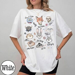 Może przedstawiać: Biała koszulka z kresk&oacute;wkowymi ilustracjami postaci z filmu Zwierzogr&oacute;d, w tym Nicka Wilde'a, Judy Hopps i Flasha. Koszulka ma napis "White" w okręgu po lewej stronie u dołu.