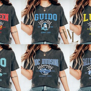 May include: Dark gray t-shirts featuring various characters from the Disney Cars movie. Each shirt displays a character's name in a collegiate font, along with a corresponding car graphic, checkered flags, and the text "Disney Cars".