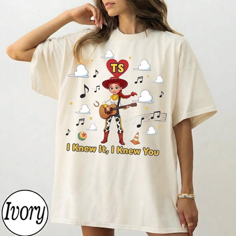 May include: An ivory t-shirt featuring a graphic of a cowgirl character playing an acoustic guitar. Above the character is a red heart with the letters TS. The design includes white clouds, musical notes, a ball, and a traffic cone with the text I Knew It, I Knew You printed in yellow below the character.