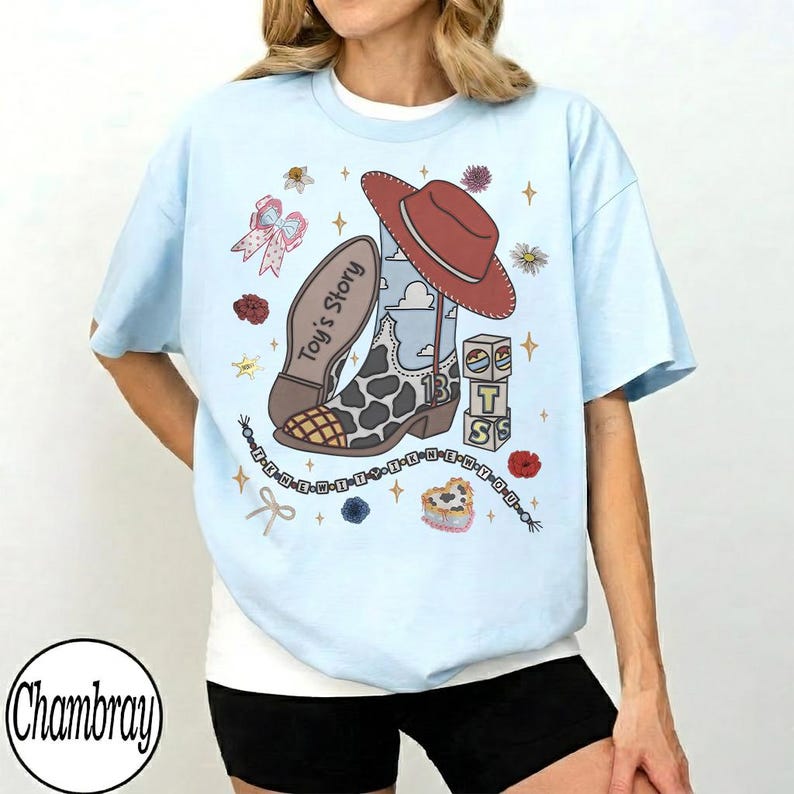 Op de afbeelding: Een lichtblauw t-shirt met een grafische print van een cowboylaars met koeienprint, een rode cowboyhoed, blokken met de letters T, S en een ster, en de tekst Toy's Story. Het ontwerp bevat kleine bloemen, sterren en een kralenketting.
