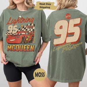 May include: Green t-shirt with a distressed Lightning McQueen graphic from the Cars film. The front shows the number 95, "Lightning McQueen," and a chequered flag. The back features a large number 95 and the character's name in a vintage style.