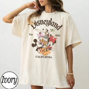 May include: Ivory-colored t-shirt featuring a Disneyland graphic with Mickey Mouse, Minnie Mouse, Donald Duck, Daisy Duck, Goofy, and Pluto. The text "Disneyland" arches across the top, with "Est 1955" and "California" below.