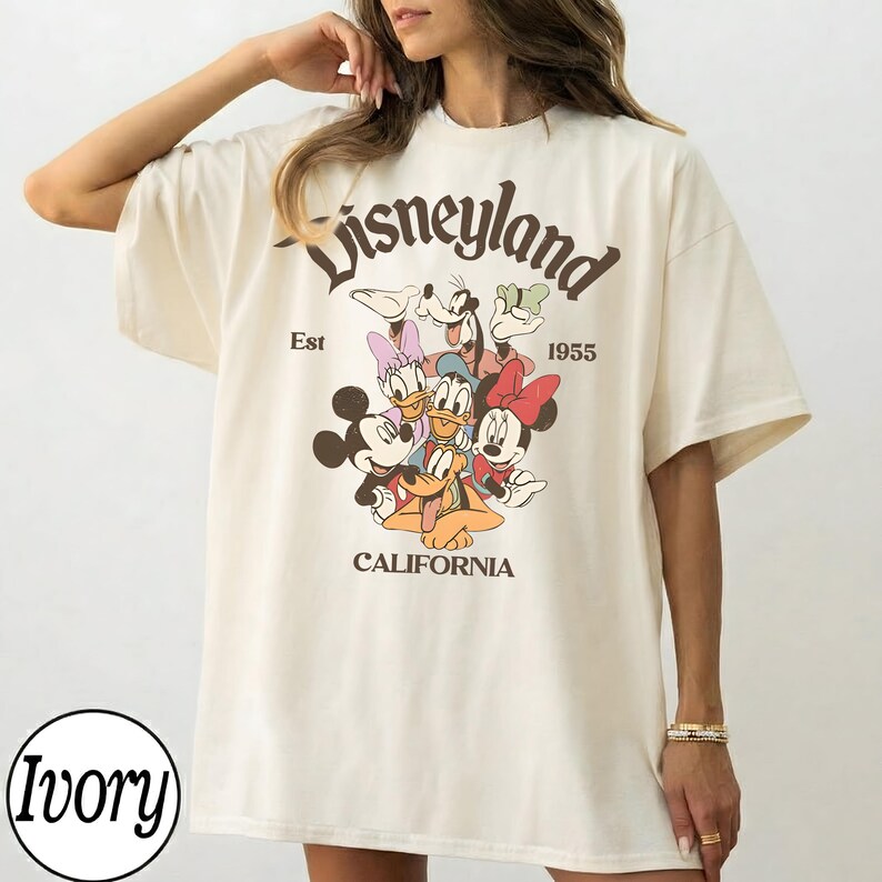 May include: Ivory-colored t-shirt featuring a Disneyland graphic with Mickey Mouse, Minnie Mouse, Donald Duck, Daisy Duck, Goofy, and Pluto. The text "Disneyland" arches across the top, with "Est 1955" and "California" below.