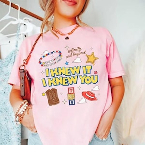 K&ouml;nnte beinhalten: Ein hellrosa T-Shirt mit einem Grafikdruck, der den Text "I KNEW IT I KNEW YOU", "infinity and beyond" und "YOU'VE GOT A FRIEND IN ME" enth&auml;lt. Das Design zeigt Illustrationen eines Perlenarmbands, eines Sterns, Cowboystiefeln, Baukl&ouml;tzen und eines Hutes.