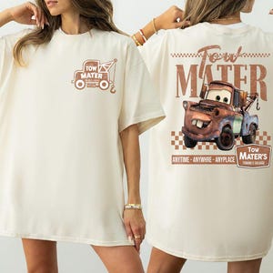May include: Cream-colored t-shirt featuring a graphic of Tow Mater from the movie Cars. The front of the shirt has a small image of Mater's tow truck, while the back displays a larger image of Mater with text that reads "Tow Mater's Towing & Salvage."