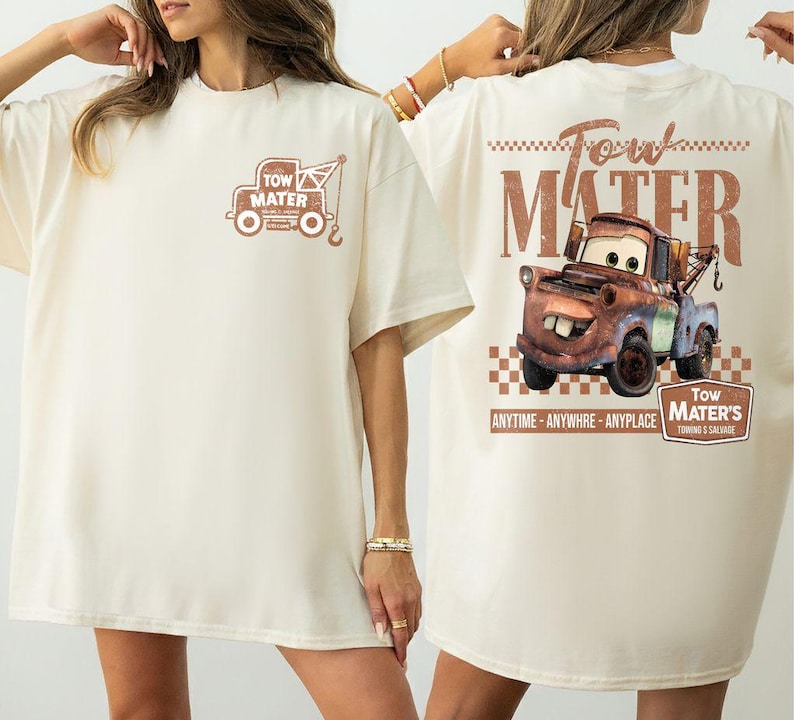 May include: Cream-colored t-shirt featuring a graphic of Tow Mater from the movie Cars. The front of the shirt has a small image of Mater's tow truck, while the back displays a larger image of Mater with text that reads "Tow Mater's Towing & Salvage."