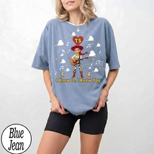 May include: A blue jean coloured t-shirt featuring a graphic of a cowgirl character playing a guitar. Above the character is a red heart with the letters TS. The text I Knew It, I Knew You is printed below the character, surrounded by clouds and musical notes.