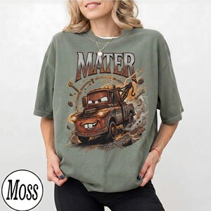 Puede incluir: Camiseta verde salvia con una imagen de dibujos animados de Mater de la pel&iacute;cula Cars. El gr&aacute;fico incluye la palabra "MATER" en una fuente desgastada, junto con rayos y herramientas. La camiseta tiene un corte holgado.
