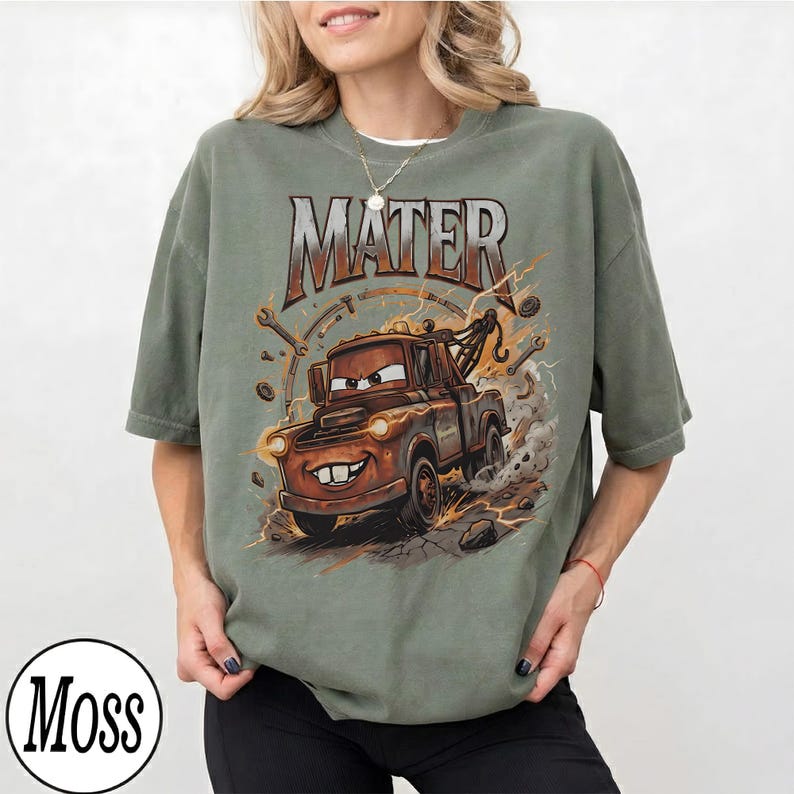 Puede incluir: Camiseta verde salvia con una imagen de dibujos animados de Mater de la pel&iacute;cula Cars. El gr&aacute;fico incluye la palabra "MATER" en una fuente desgastada, junto con rayos y herramientas. La camiseta tiene un corte holgado.