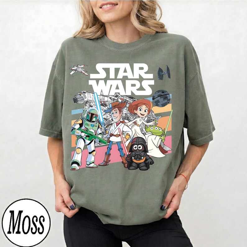May include: Olive green t-shirt with a Star Wars graphic featuring characters including Darth Vader and Woody. The Star Wars logo is above the characters. The shirt has a loose fit and is made of soft material.