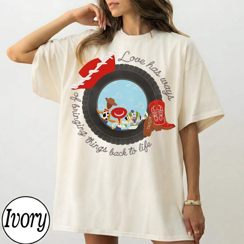 Op de afbeelding: Een ivoorkleurig t-shirt met een afbeelding van een zwarte band die een sc&egrave;ne omlijst met een cowboyhoed, een rode cowboylaars en stripfiguren. De tekst luidt Love has ways of bringing things back to life, met Taylor op de laars.