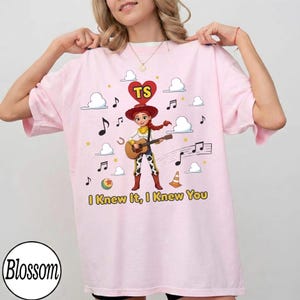 May include: A light pink t-shirt featuring a graphic of a cowgirl character playing an acoustic guitar. Above the character is a red heart with the letters TS. The design includes musical notes, clouds, a ball, and a traffic cone, with the text I Knew It, I Knew You printed in yellow at the bottom.