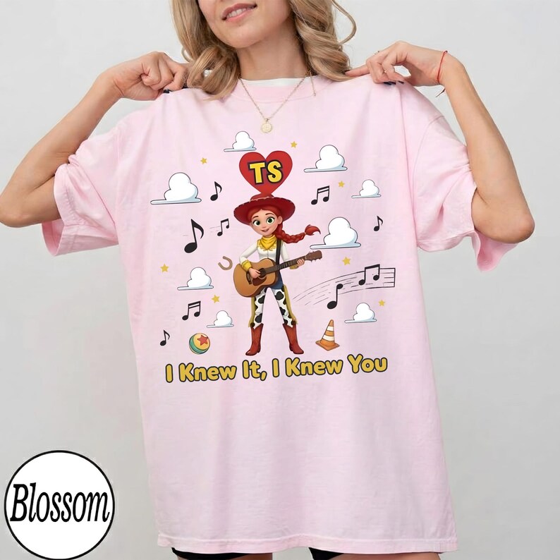 May include: A light pink t-shirt featuring a graphic of a cowgirl character playing an acoustic guitar. Above the character is a red heart with the letters TS. The design includes musical notes, clouds, a ball, and a traffic cone, with the text I Knew It, I Knew You printed in yellow at the bottom.