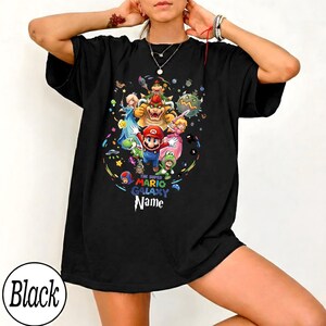 May include: Black t-shirt with a vibrant graphic of Mario and friends from Super Mario Galaxy. The text "The Super Mario Galaxy Name" is printed below the characters. The shirt is a relaxed, oversized fit.