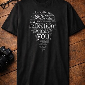 May include: Black t-shirt with a white text design. The text is arranged in a shape and includes the words "Everything", "see", "reflection", and "within you". The shirt is displayed on a wooden surface.