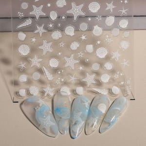 May include: Clear nail art stickers featuring white starfish, seashells, and other marine life designs. The stickers are applied to several fingernails with a blue and white marbled effect.