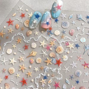 May include: A sheet of nail art stickers featuring various seashells and starfish in shades of blue, pink, and white, alongside three painted nail tips displaying a gradient design with small starfish and shell embellishments.