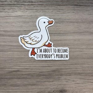 Può includere: Un adesivo raffigurante un'anatra bianca in stile cartone animato con piedi e becco arancioni, accompagnato dal testo nero I'M ABOUT TO BECOME EVERYBODY'S PROBLEM. Il disegno presenta un bordo bianco ed è realizzato con linee semplici e nette.