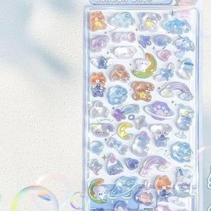 Puede incluir: Una lámina de pegatinas transparentes e iridiscentes con varios diseños bonitos, incluyendo osos, nubes, estrellas y arcoíris. Las pegatinas están en un paquete de plástico con el texto "BONBON DROP".