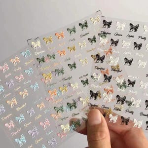 May include: Nail art stickers with small, textured bows in pastel shades. Each bow has a word like "Pretty" or "Grace" next to it. The stickers are on a clear backing, with the text "MN-102F" visible.