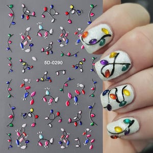 May include: A sheet of 5D nail art stickers featuring various designs of colorful Christmas light bulbs on black wires, labeled 5D-0290. The stickers are shown applied to white glittery fingernails, displaying individual light bulb patterns.