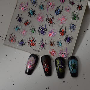 Könnte beinhalten: Ein Bogen mit selbstklebenden Nagelstickern, die verschiedene Spinnenmotive in mehreren Farben wie Rot, Lila, Blau und Grün mit Glitzerakzenten zeigen, beschriftet mit Co-504. Vier schwarze künstliche Fingernägel zeigen aufgebrachte Spinnensticker.