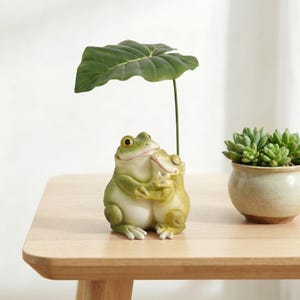 May include: A whimsical frog figurine in shades of green, embracing another frog. A large green leaf is held above the frogs. A small potted succulent sits to the right. The figurine is on a light-colored wooden surface.