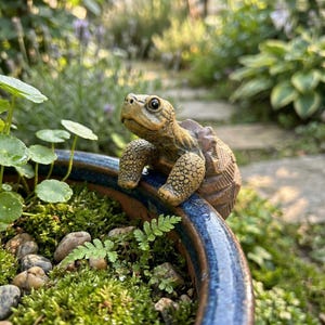 Puede incluir: Una detallada figura de tortuga marrón posada en el borde de una maceta de cerámica azul y marrón. La tortuga tiene la piel texturizada y está rodeada de vegetación, incluyendo musgo, pequeñas rocas y helechos. El fondo está borroso.