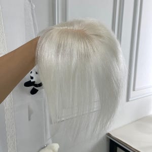 May include: A hair topper featuring straight, bright white hair with a visible scalp-coloured base at the crown. The hair is smooth and falls in a layered style, showcasing a clean part line and a natural-looking transition from the base to the strands.