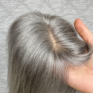 Puede incluir: Un postizo de cabello liso en tono gris plateado con una raya de color piel de aspecto natural. El cabello es fino y suave, mostrando una mezcla de tonos grises claros y oscuros que imitan el cabello natural con una base realista.