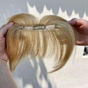 Puede incluir: Una pieza de cabello rubio claro con una base de malla y clips metálicos. La pieza de cabello está sostenida por las manos de una persona, mostrando el color y la textura del cabello. Diseñada para añadir volumen o longitud.