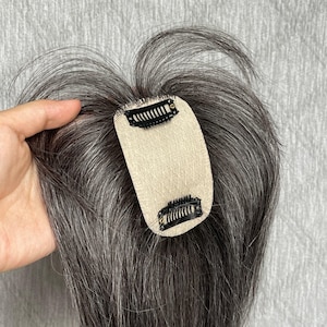 May include: A hair topper featuring straight, salt-and-pepper gray strands attached to a beige, oval-shaped mesh base. Two black metal pressure-sensitive clips are secured to the top and bottom of the mesh, designed for attachment to natural hair.