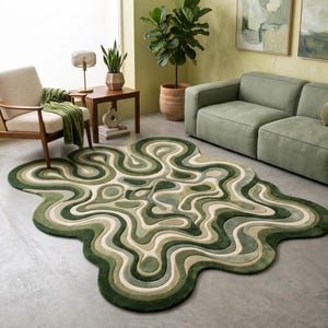 Może przedstawiać: Abstrakcyjny dywan w odcieniach zieleni i złamanej bieli, o falistym, organicznym wzorze. Dywan ma różne odcienie zieleni, tworząc efekt warstwowy. Dywan leży na betonowej podłodze, w tle meble i rośliny.