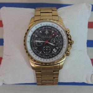 Könnte beinhalten: Goldfarbene Armbanduhr mit schwarzem Zifferblatt und weißem Außenring. Die Uhr verfügt über mehrere Hilfszifferblätter, rote Zeiger und den Text "SPIRIT OF CONCORDE". Die Uhr hat ein goldfarbenes Metallarmband.