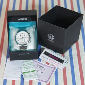 May include: A silver-toned analog wristwatch with a white dial and multiple sub-dials, presented in a black box with a light blue interior. The box lid features the WIRED logo. Included are a white instruction booklet and a warranty card.