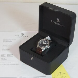 May include: A silver-tone analog wristwatch with a black dial, white Arabic numerals, and a brown leather strap, presented inside a black rectangular box with the Steinhart logo printed in white on the interior lid and a document nearby.