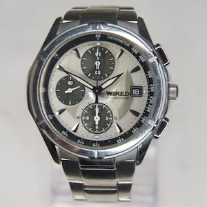 May include: A silver-toned stainless steel Wired chronograph watch featuring a metallic silver dial with three dark gray sub-dials, a date window at the three o'clock position, and a black tachymeter bezel. The watch has a link bracelet band.