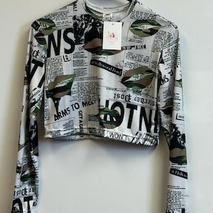 May include: A white long-sleeved shirt with a cropped length. The shirt has a print of black and white text and green camouflage patterns. The text includes the words "HOTNI", "WON'T", "ARMS TO MELT", and "GET A GRE!"
