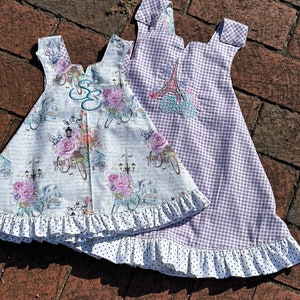 May include: Two sleeveless dresses with white ruffles and black polka dot trim. One dress has a floral print with a bicycle and streetlights. The other dress is purple and white gingham with an embroidered Eiffel Tower and bicycle.