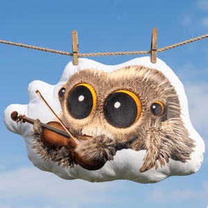 May include: A whimsical, cloud-shaped pillow featuring a large-eyed, furry spider playing a miniature violin. The pillow is primarily white with brown and tan details, and is suspended from a clothesline against a blue sky.