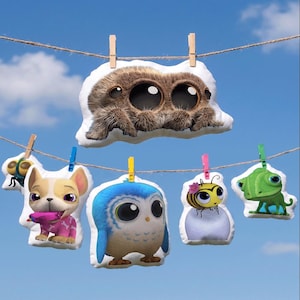 Puede incluir: Una colección de cojines de peluche con personajes, incluyendo una araña, un perro, un búho, una abeja y un lagarto, colgados de una cuerda de tender la ropa contra un cielo azul. Cada cojín tiene un diseño único y colores vibrantes.